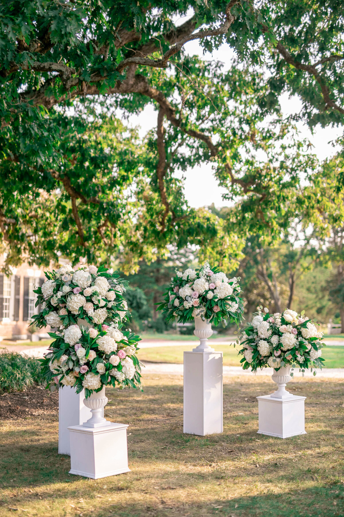 Three white floral arrangements on pedestal stands are set outdoors under a large leafy tree. Each arrangement features lush white and pink flowers with greenery, creating an elegant and serene atmosphere in a garden setting.