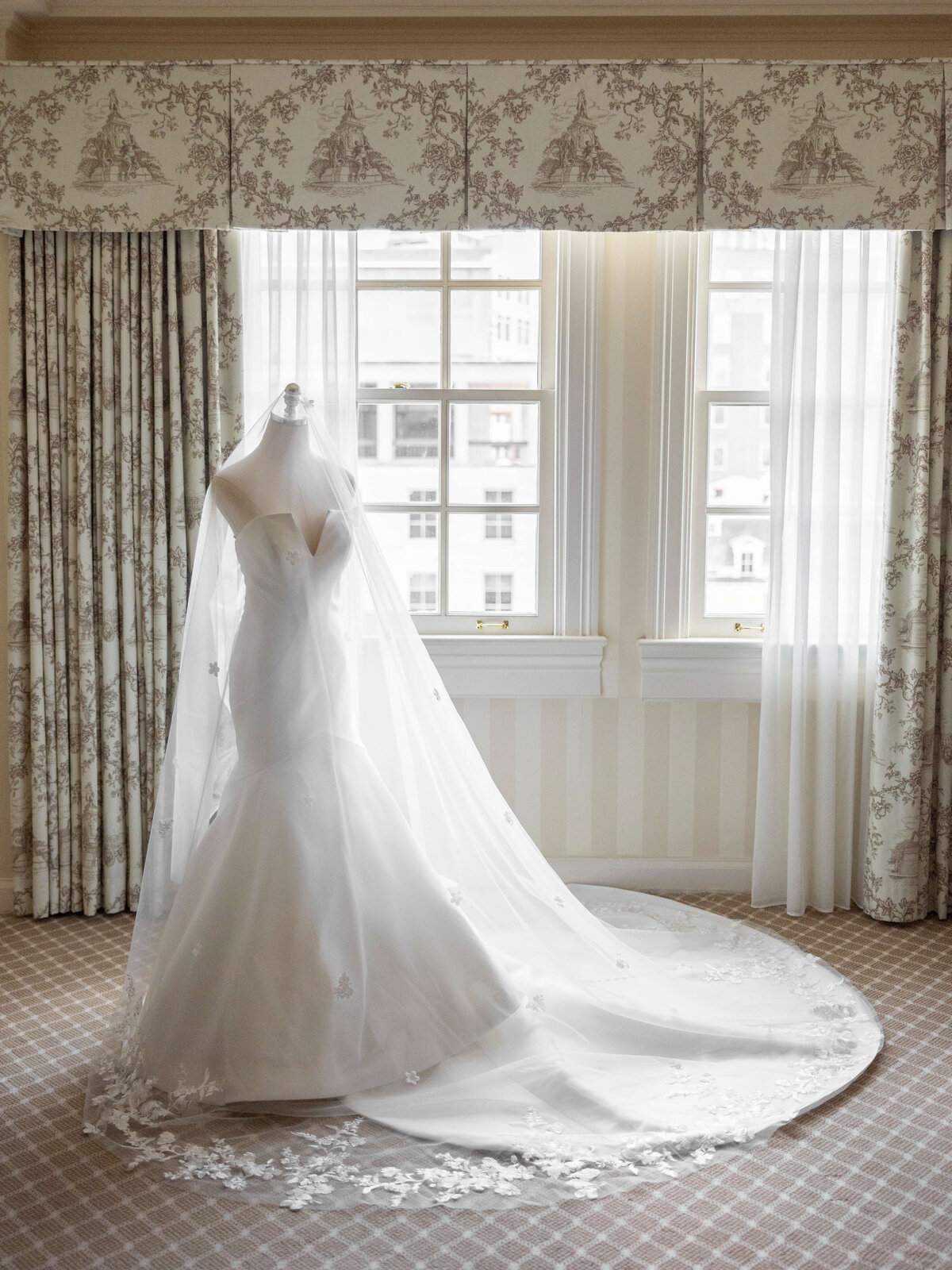 A white strapless wedding gown on a mannequin, draped with a long, embroidered veil, is positioned in front of a softly lit window. Floral-patterned curtains frame the window, and the room is carpeted with a subtle checked design.