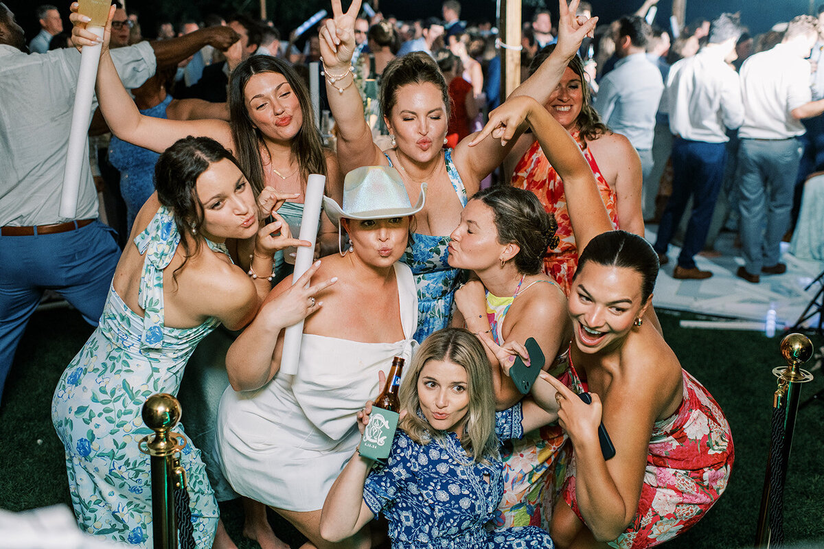 Bride and friends posing with glow sticks and drinks during a lively dance floor party under a sailcloth tent.