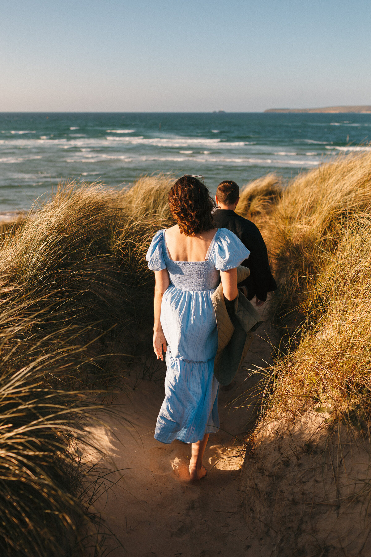 Couple walking down the beach the girl wears a blue dress which catches the wind, the sea is blue behind as they descend the sand dunes