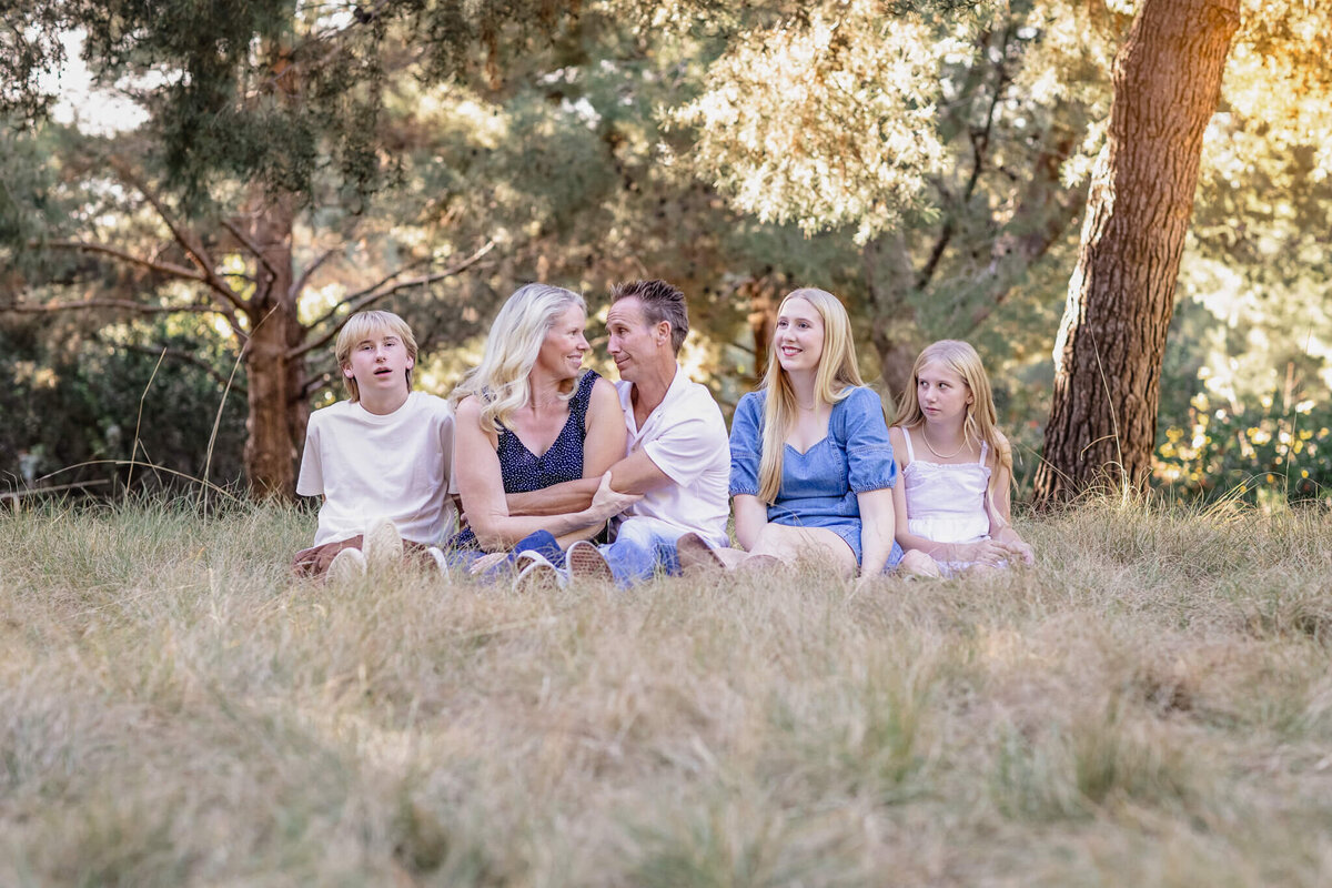 A family of five with blonde hair sitting in the grass next to each other, looking at one another and laughing and smiling at Jeffrey Open Space.