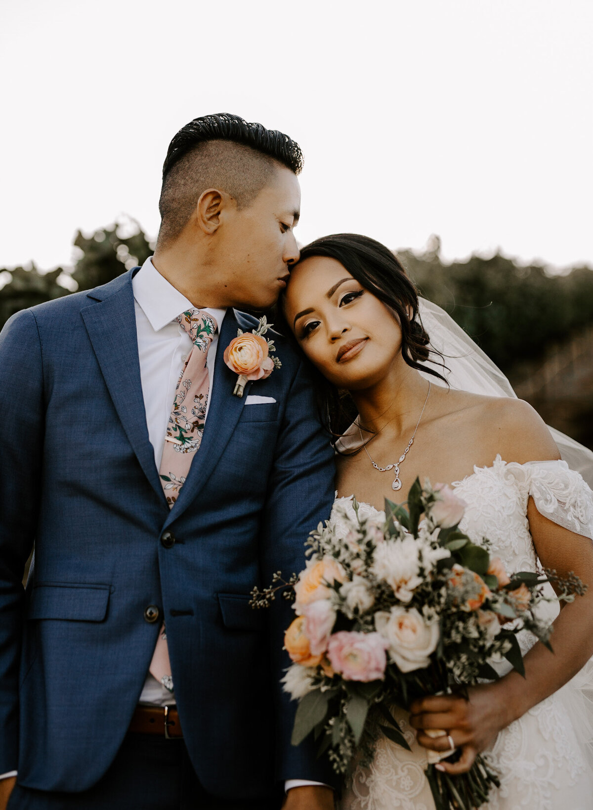 Groom kisses his brides head while holding a bouquet of wedding flowers