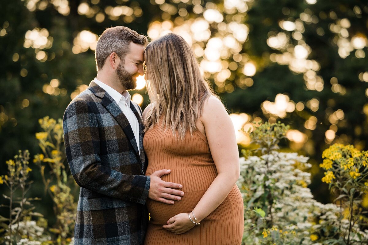 A couple embracing affectionately, with the man's hand on the woman's pregnant belly, against a blurred natural backdrop bathed in warm sunlight. This moment captured by a skilled Pittsburgh maternity photographer creates a