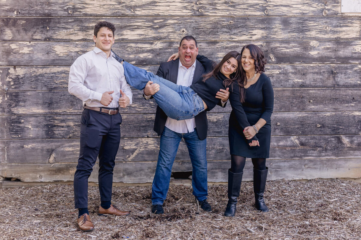 A playful family of four smiling and laughing as son puts two thumbs up, dad lifts up daughter laughing, and mom puts head against daughter's and smiles.