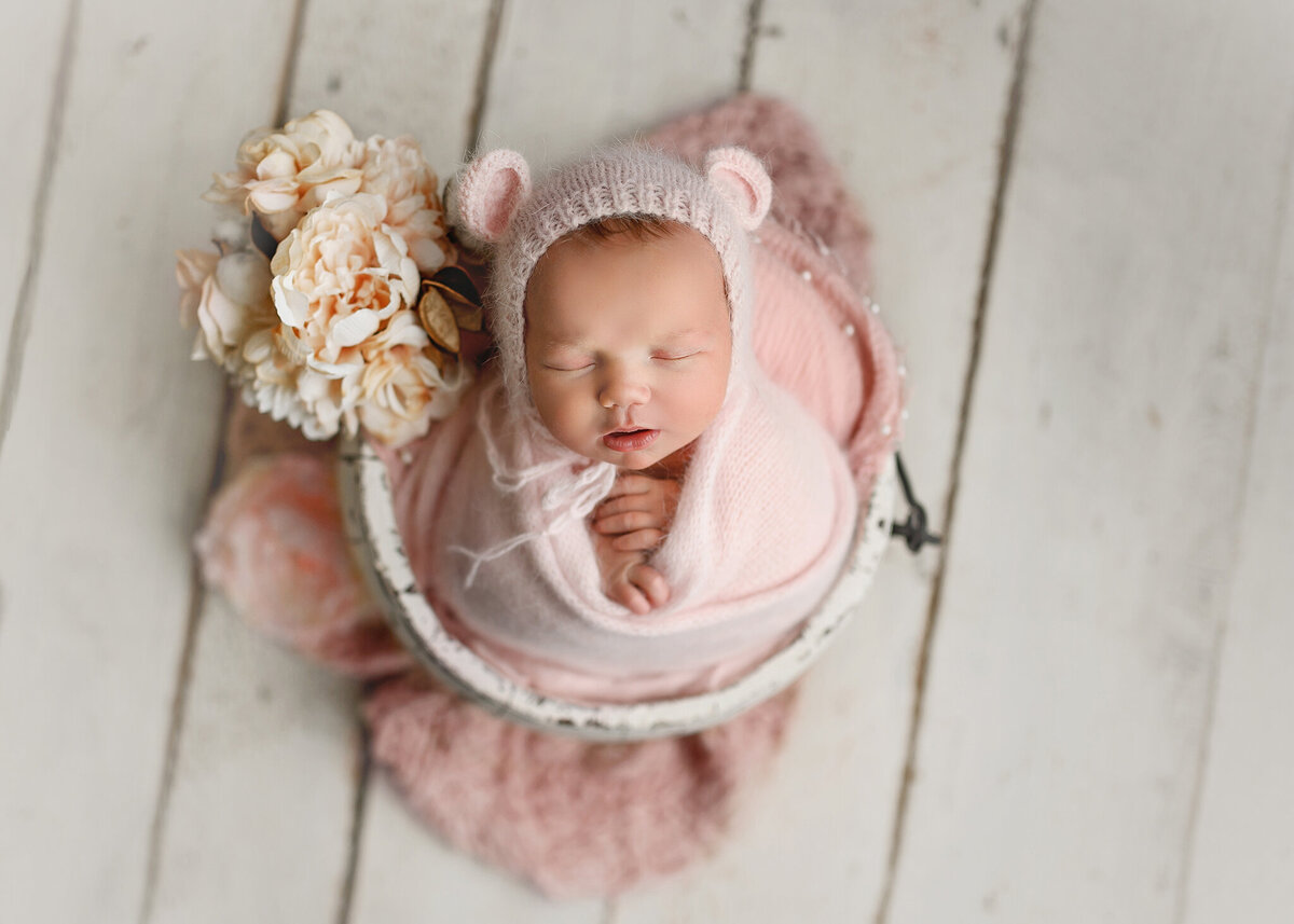 newborn girl wearing a pink swaddle and bear bonnet posed in a white bucket with at her side