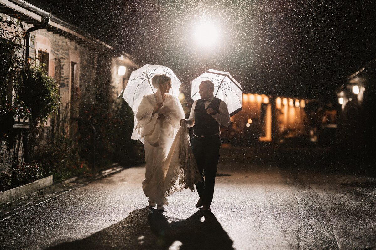 Bride and groom walking under umbrellas at night in the rain outside Loch Lomond Arms Hotel in Luss, Scotland, sharing a romantic and elegant wedding moment.