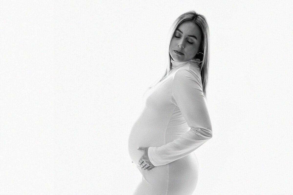 A pregnant woan in a white maternity gown with long sleeves glances over her shoulder in a studio