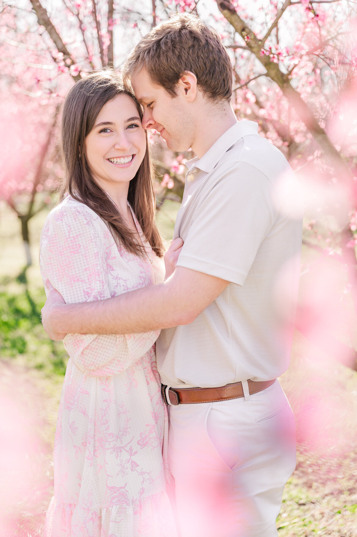A smiling and happy engaged couple wrapped in bright pink cherry blossom trees enjoying their Raleigh wedding photo session by JoLynn Photography