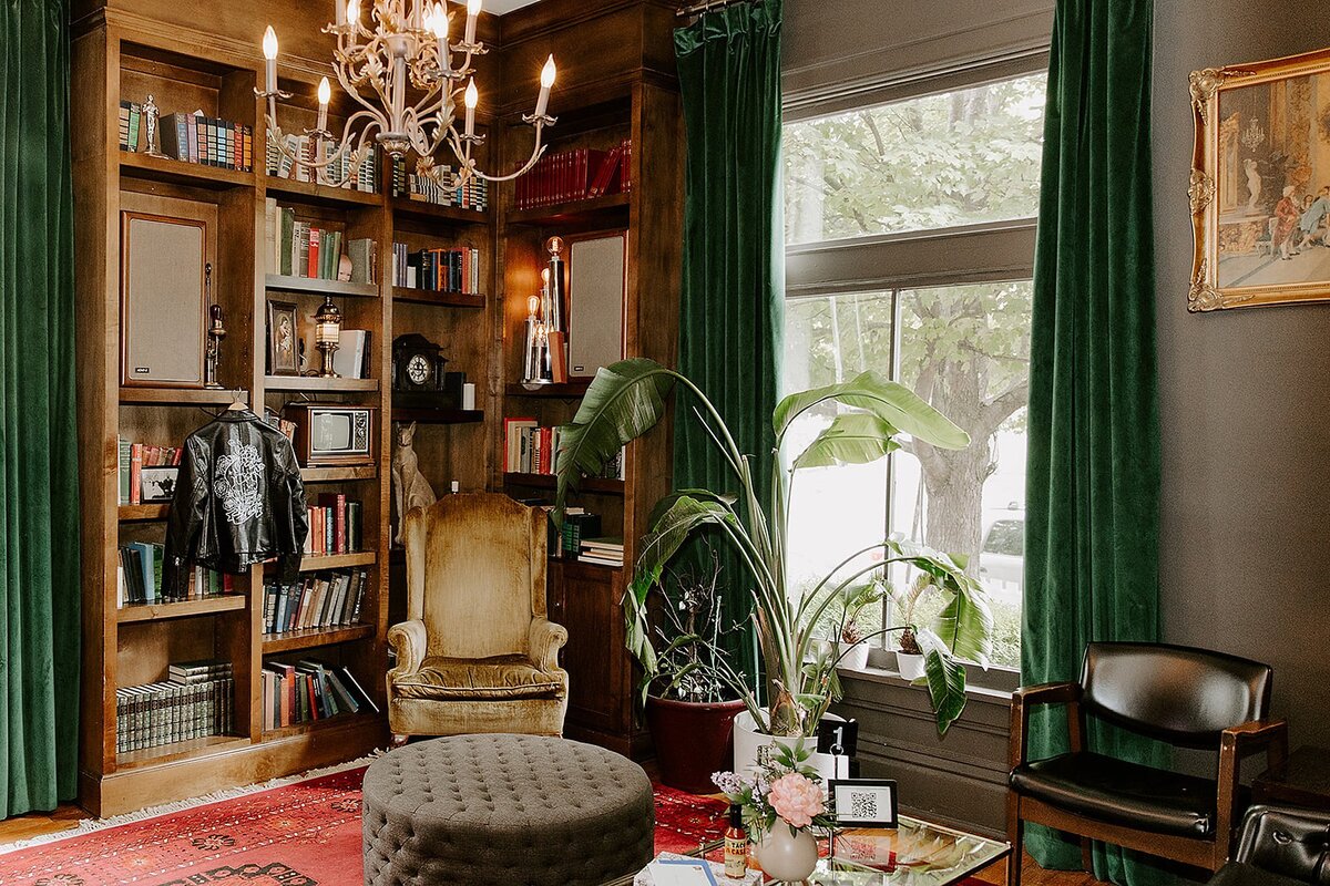 A room with tall windows and leafy monstera plants at Rosemary and Beauty Queen. On the left side of the room is a floor to ceiling bookcase with vintage books, a small TV and a speaker. The bride's black leather jacket hangs from the bookcase. A brass chandelier hangs from the ceiling above a gold velvet chair and a beige tufted ottoman on a red oriental carpet. A low brown leather chair sits off to the right under a paining in a gold frame.