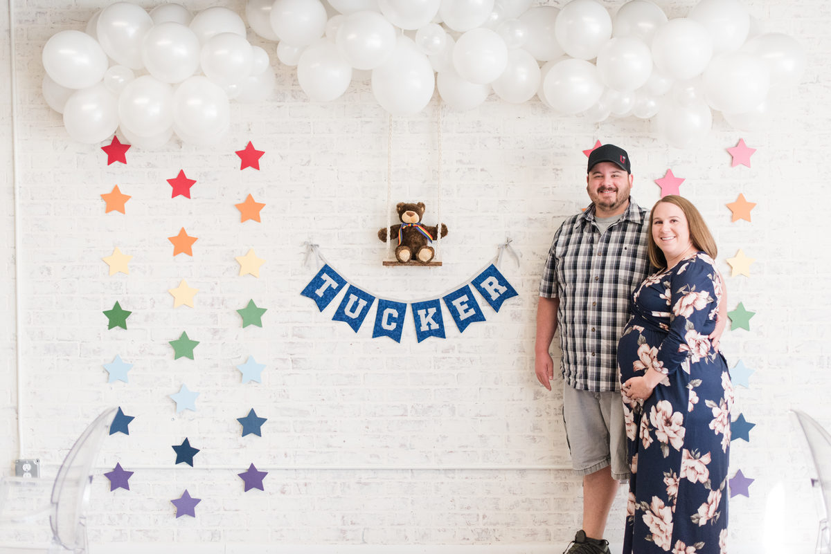 Parents-to-be standing in front of a whimsical baby boy shower setup featuring rainbow star garland, white balloon clouds, and a teddy bear on a wooden swing against a white brick backdrop.