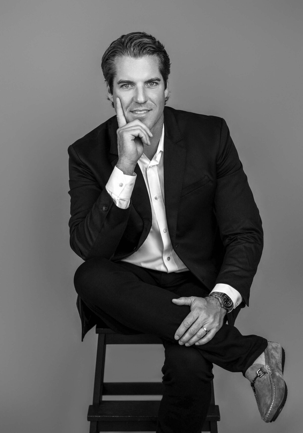 A man dressed in a black suit, sitting in a modern white chair, smiling confidently.