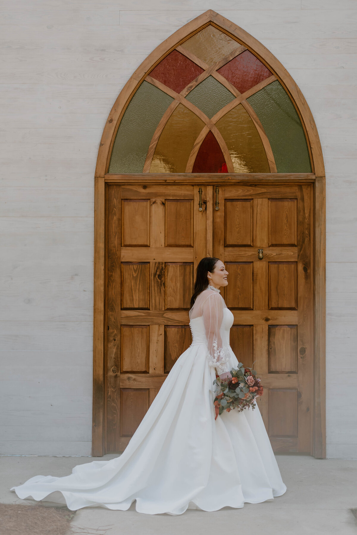a bride in her wedding dress holding her bouquet standing in front of a wood and stained glass door at cypress grove open air chapel in louisiana.