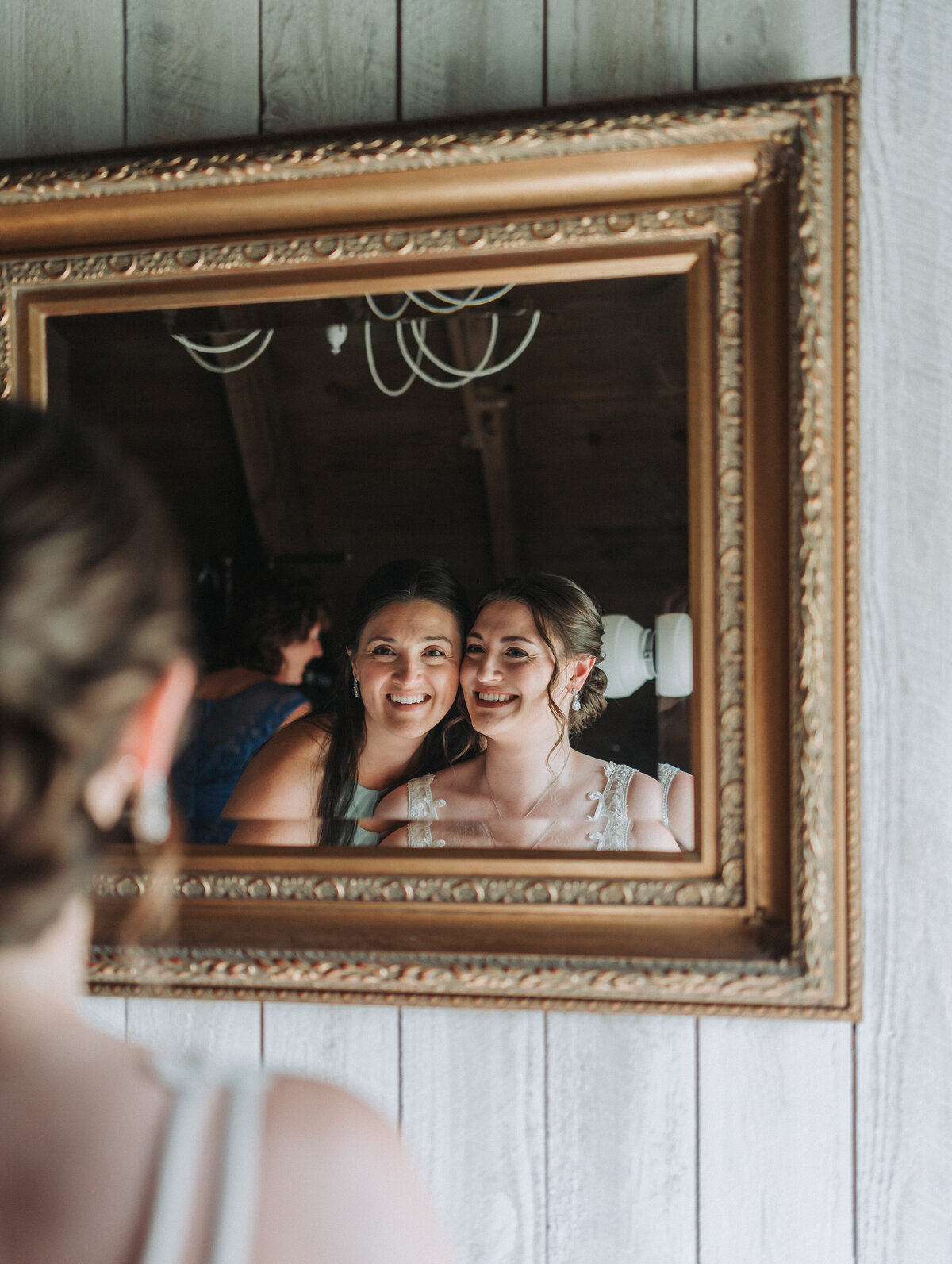 Bride and bridesmaid smile into a vintage mirror during the getting ready moments at Sweeney Barn in Manassas, Virginia.