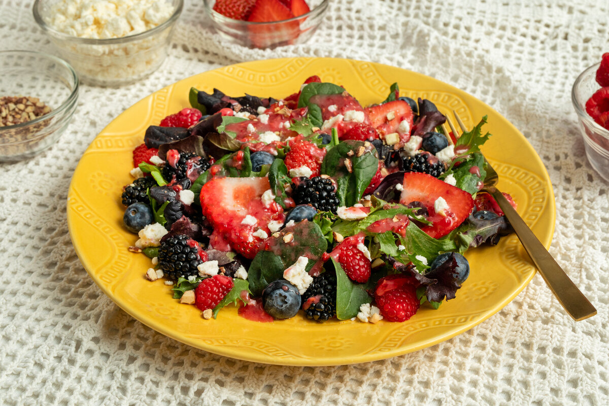 A spring berry salad with homemade raspberry vinaigrette on a yellow plate with a bowl of feta cheese and raspberries on the side.
