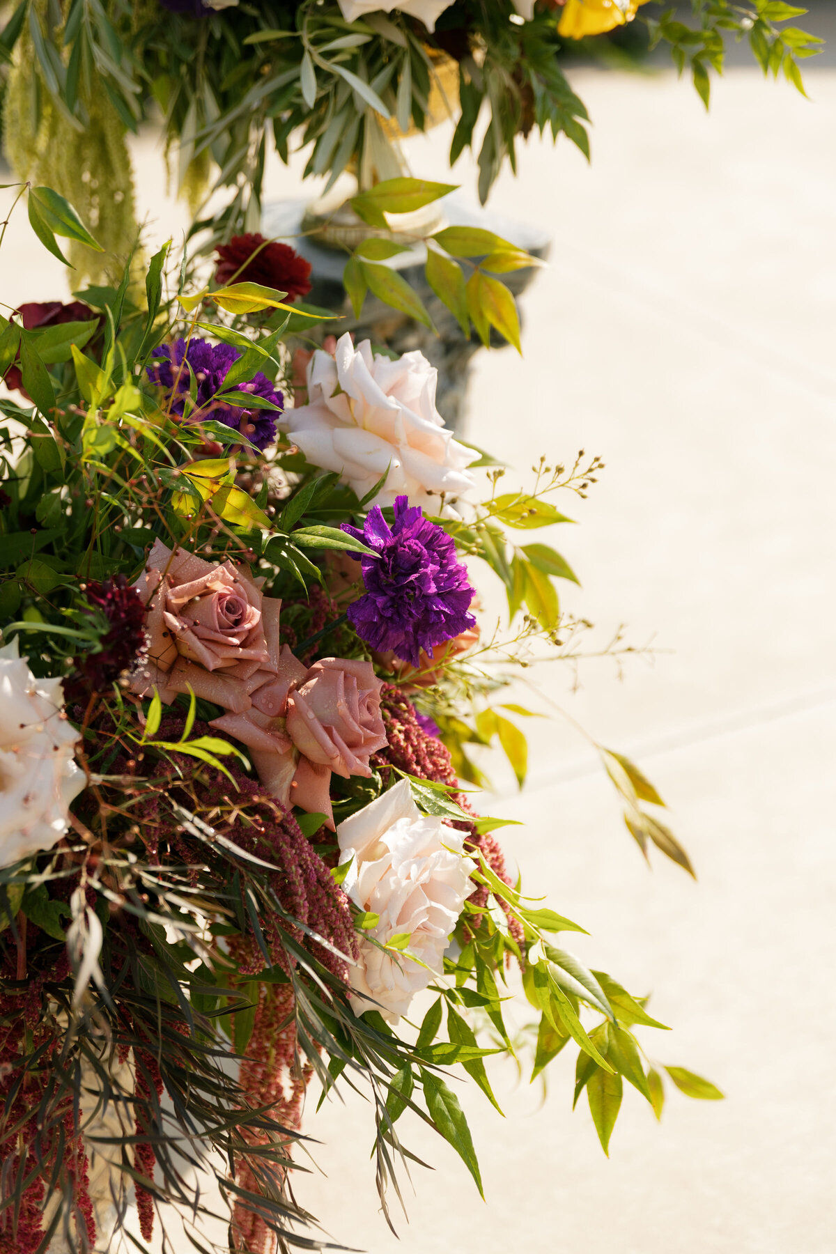 Soft, romantic floral detail of blush roses, purple blooms, and fresh greenery arranged for an outdoor wedding ceremony in natural sunlight.