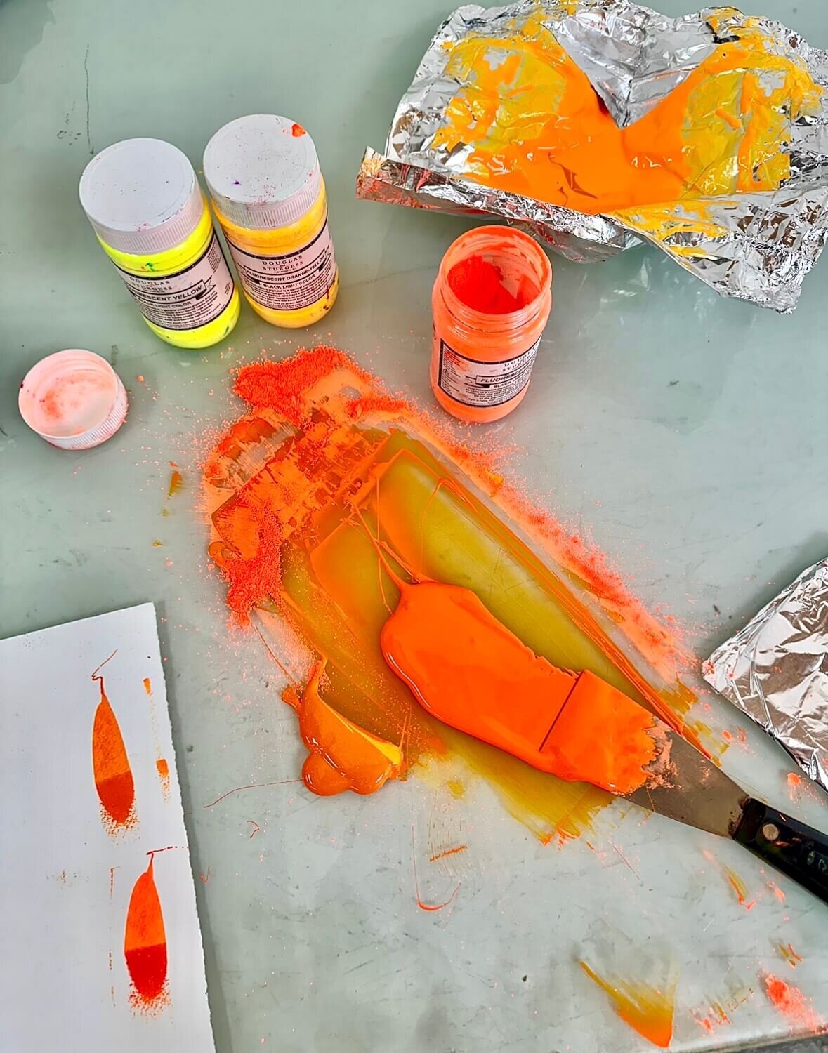 Fluorescent orange and yellow pigment powders being mixed on foil and glass, capturing the glow and texture of hand-mixed ink.