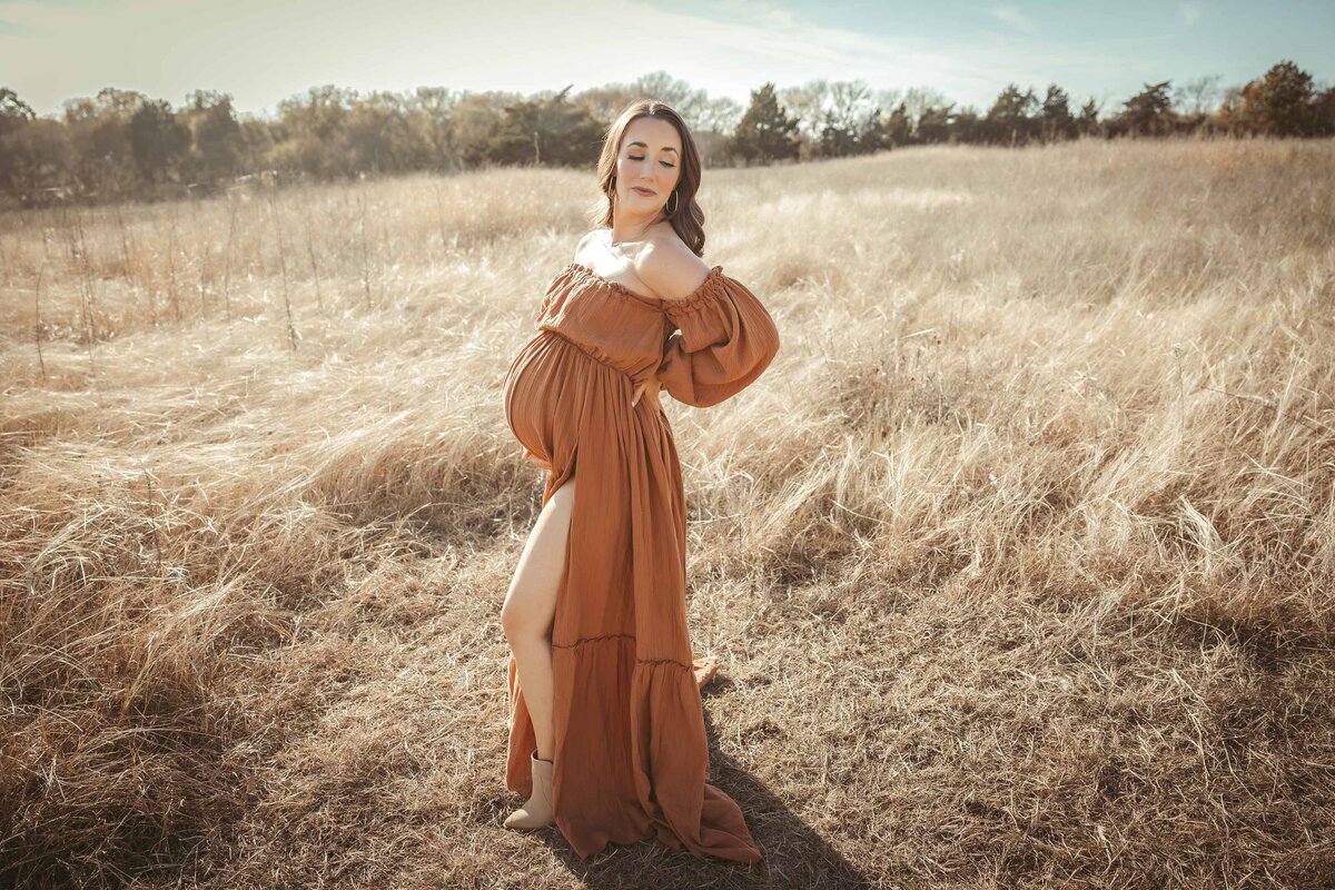 Woman in maternity session wearing a rust color dress looking over her shoulder standing in a field of grass.