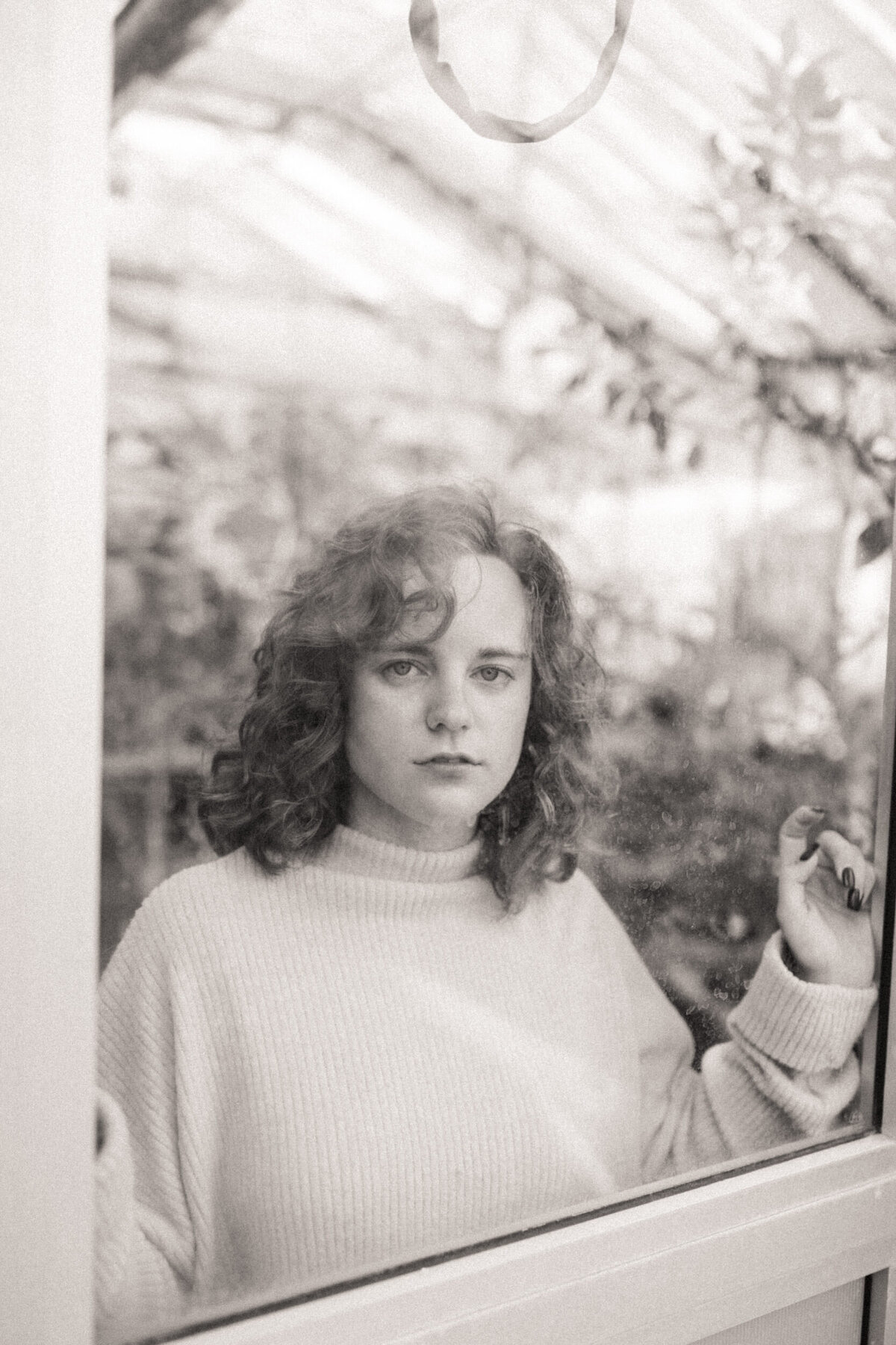 Senior with natural curly hair wearing a sweater and looking through the window in a greenhouse