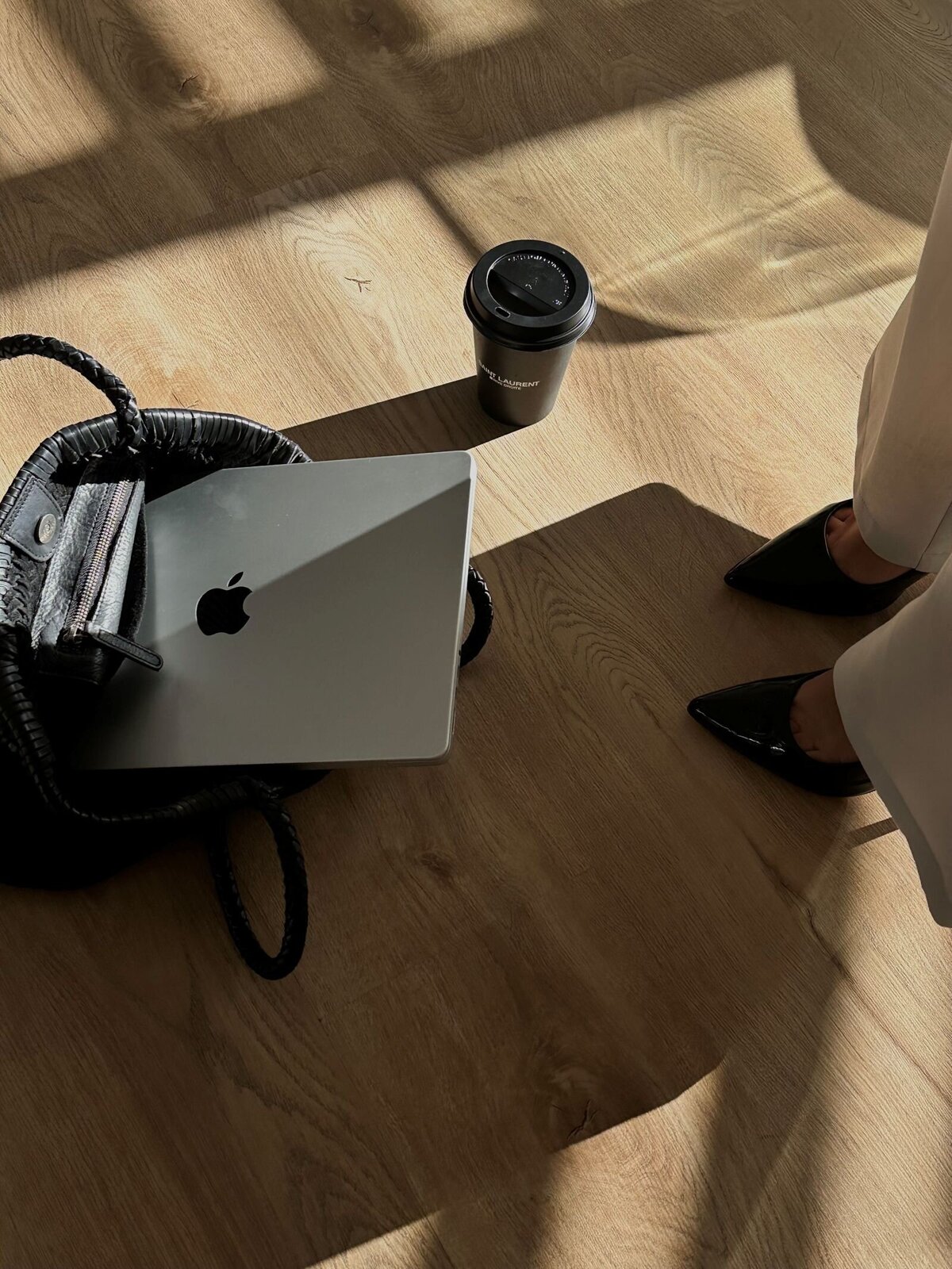 A businesswoman in cream linen pants and patent black pointy-toed stilettos stands beside her black handbag on the floor, which contains a laptop. A Saint Laurent coffee cup rests beside the handbag, completing the sophisticated, professional setting.