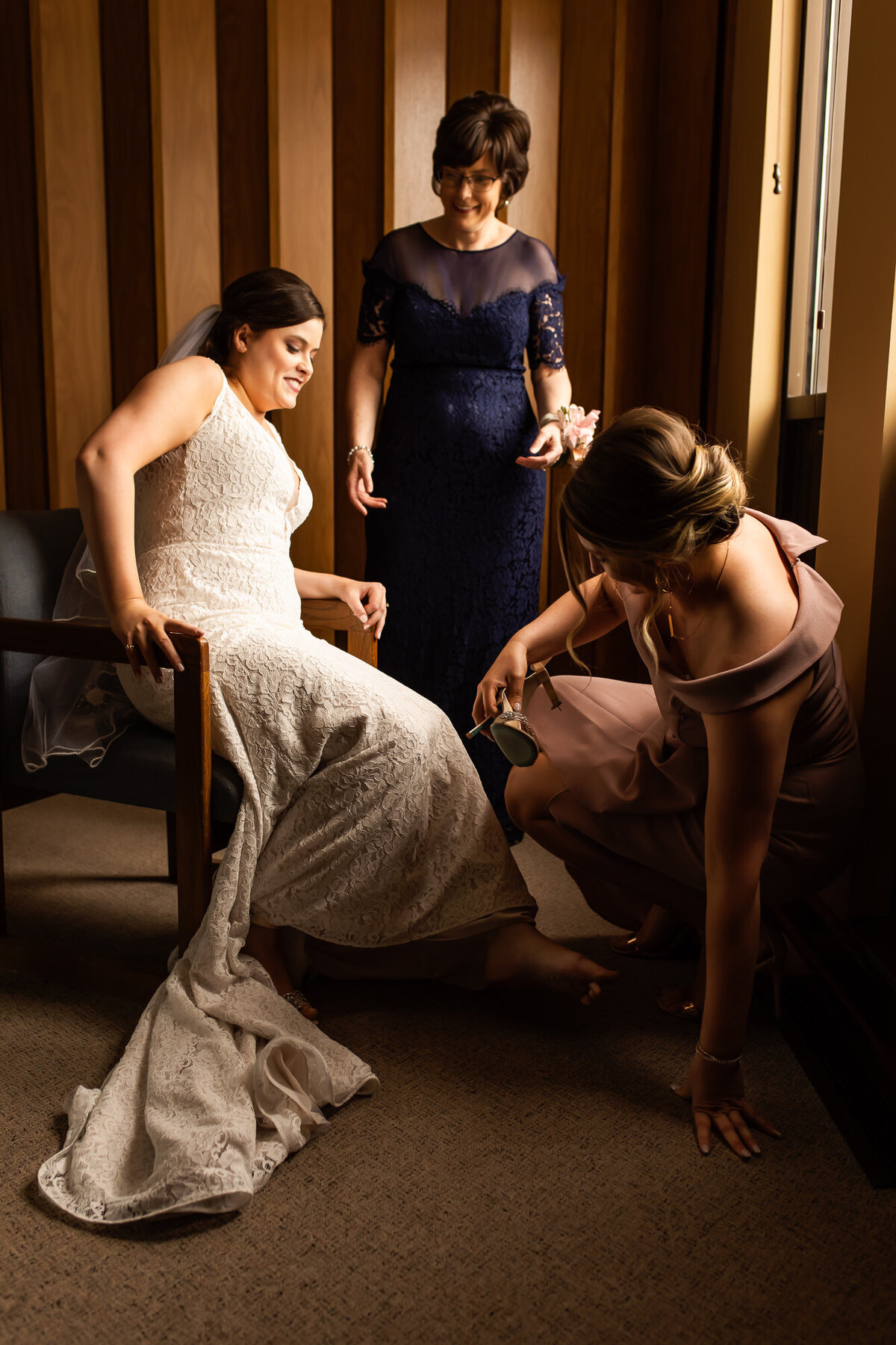 Bride sits as her maid of honor puts her shoes on her feet