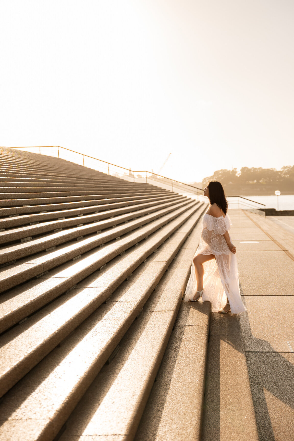 Pregnant woman in a sheer white dress walking up the iconic Sydney Opera House steps during a golden hour maternity photoshoot