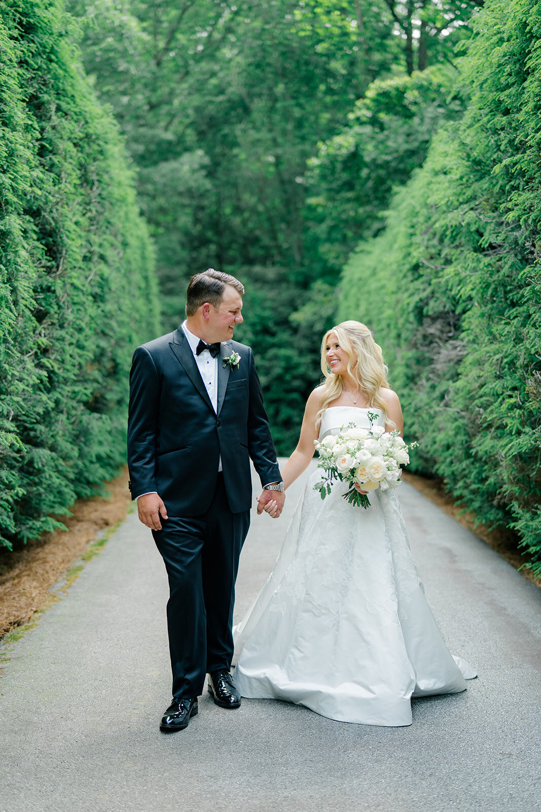  Bride and groom walking on a tree-lined path, surrounded by lush greenery at their Highlands, NC wedding.