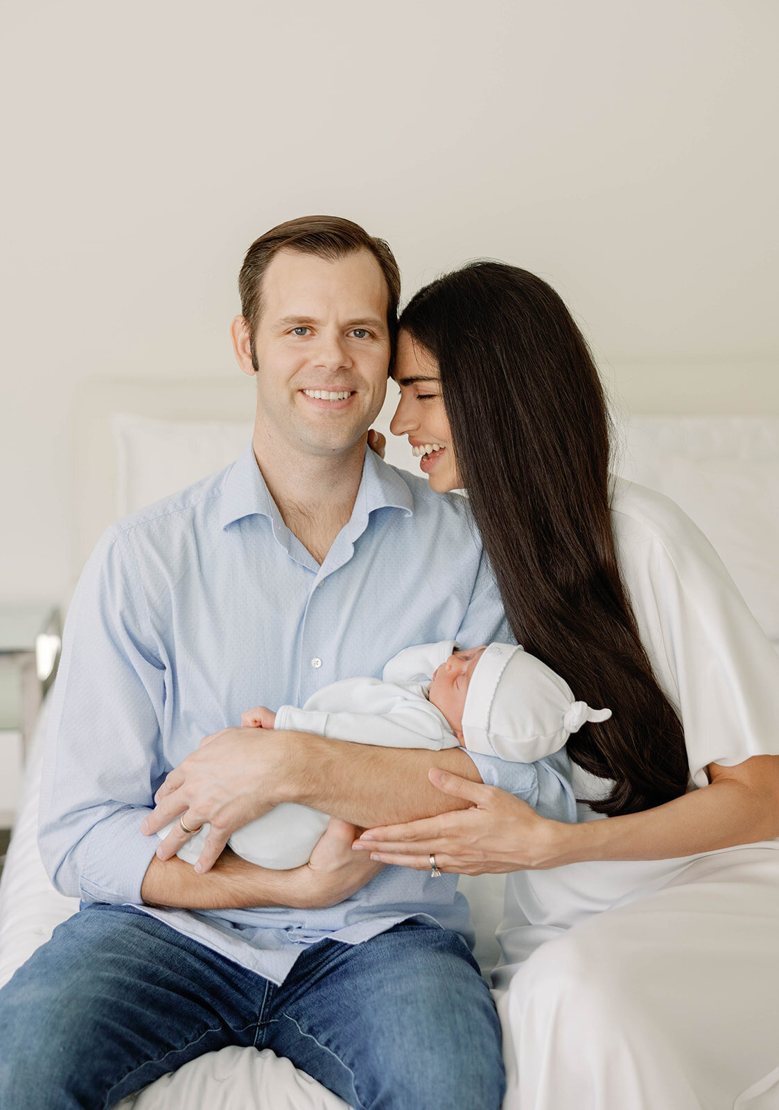 A happy couple sits on their bed, lovingly holding their newborn during a Florida Keys family photography session with Claudia Amalia.