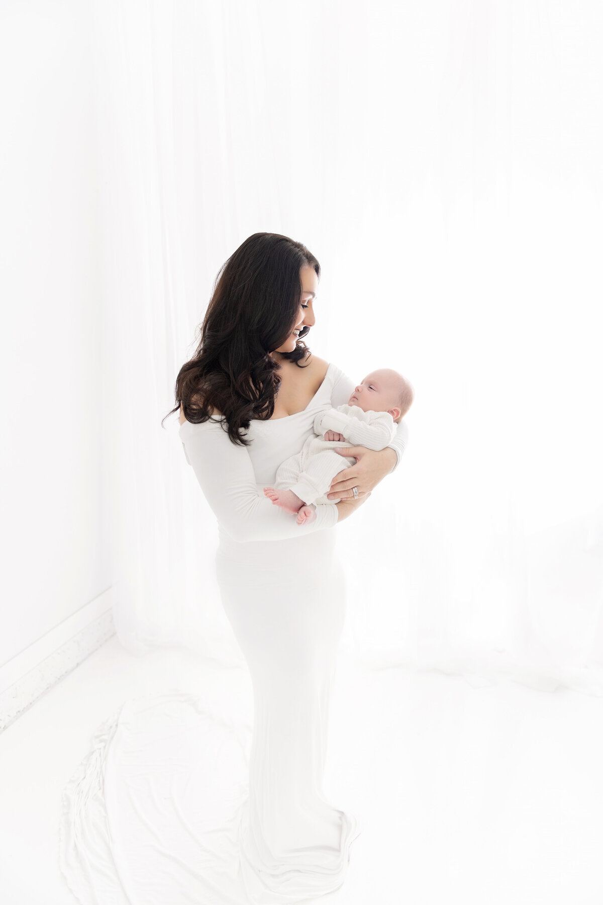A happy new mother in a glamorous white dress cradles her newborn baby while standing in a studio