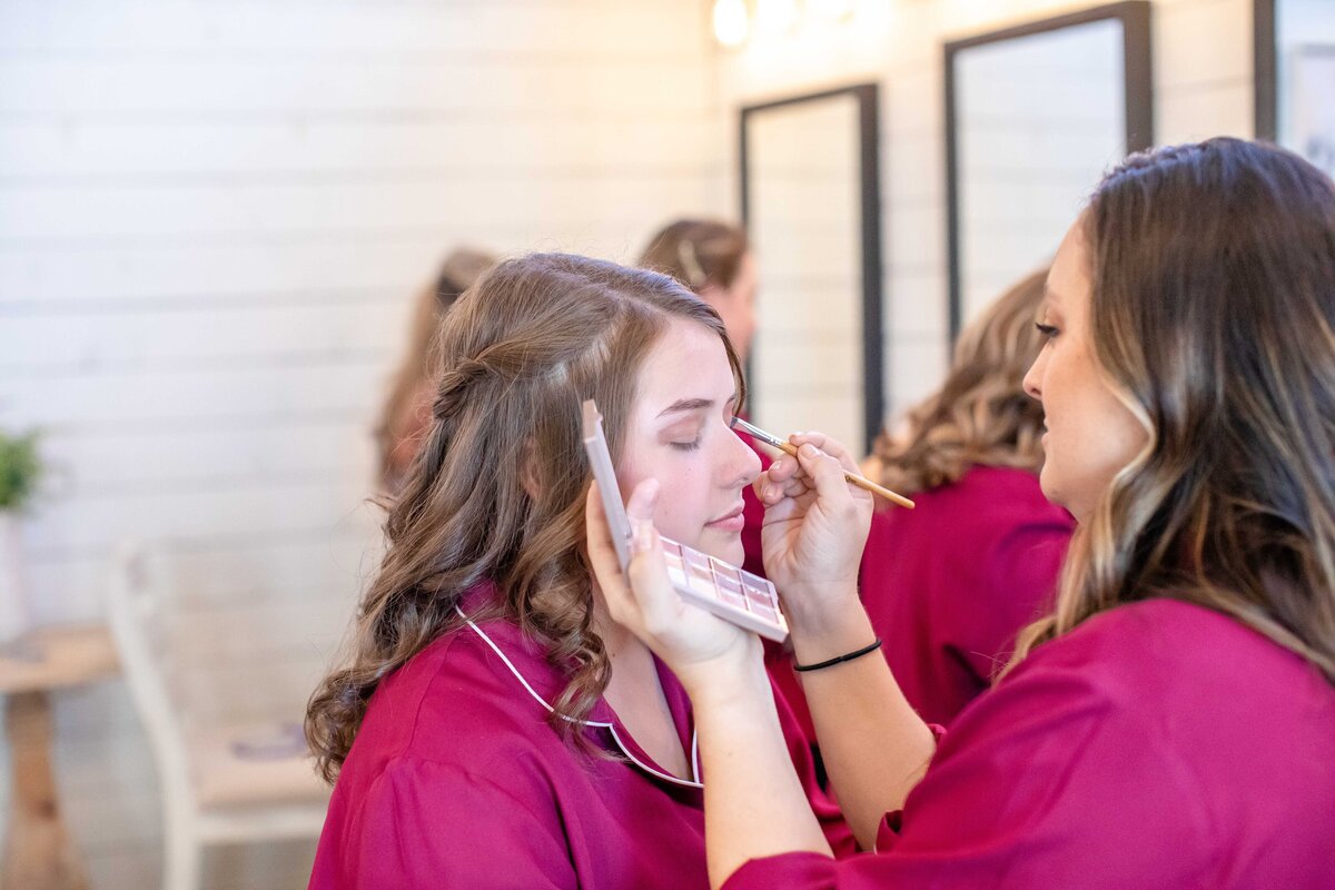 bridesmaid in maroon robe applies makeup at Morgan Creek Barn in Dripping Springs Texas wedding