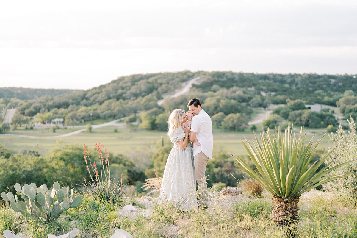 A loving family poses with their newborn in a picturesque Texas outdoor setting. The parents gaze adoringly at their baby, wrapped in a soft blanket, while warm sunlight filters through tall trees. The golden fields and sprawling landscape create a serene and heartfelt moment, capturing the joy of new life.
