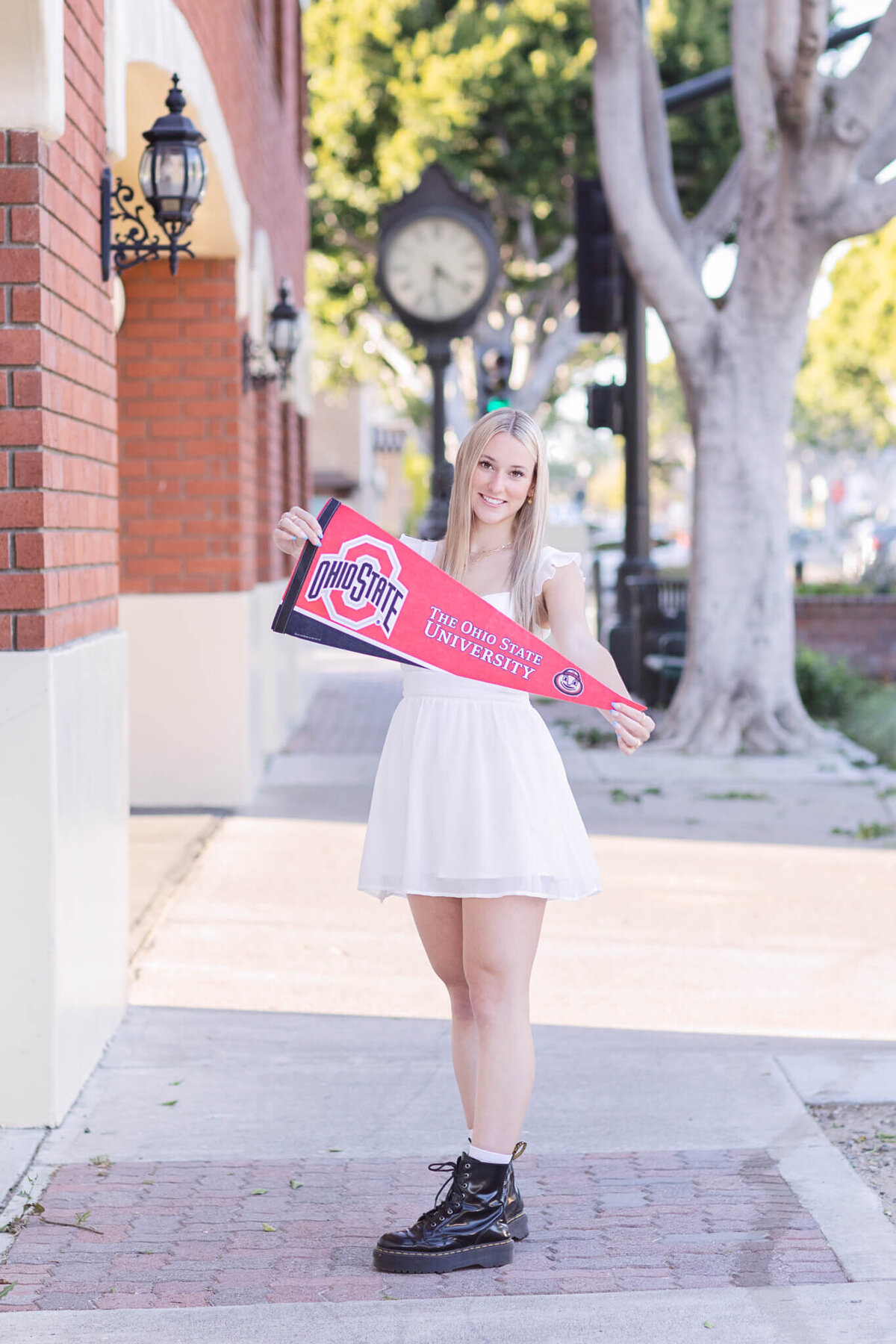 A young woman in a white dress smiles while holding an Ohio State University pennant. She stands on a sidewalk lined with trees and red brick buildings, with decorative lanterns on the wall behind her.