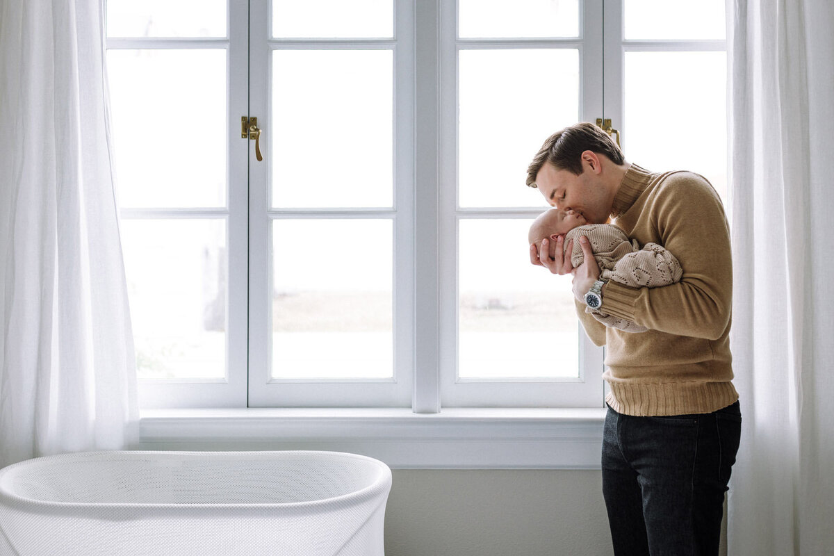 dad standing in front of large window with white curtains holding swaddled baby next to his face and kissing him