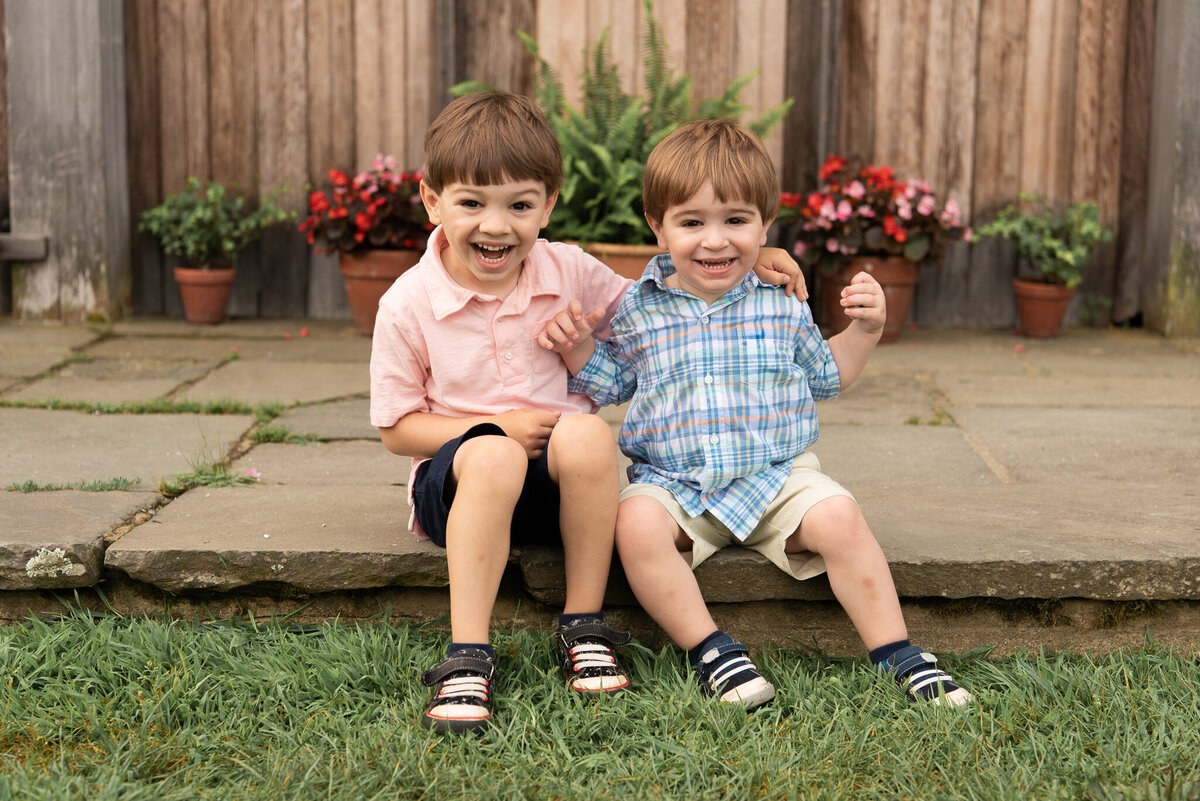 Two young boys are sitting on a step, smiling at the camera in front of a wooden wall