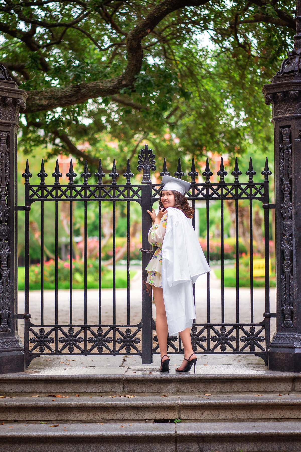 Hispanic high school senior female with white cap and gown at the gates of Jackson Square.  She is wearing black high heeled shoes and has turned her head around to face the camera.  She is holding her gown over her shoulder and is wearing her cap.  She is holding on to the wrought iron gate.