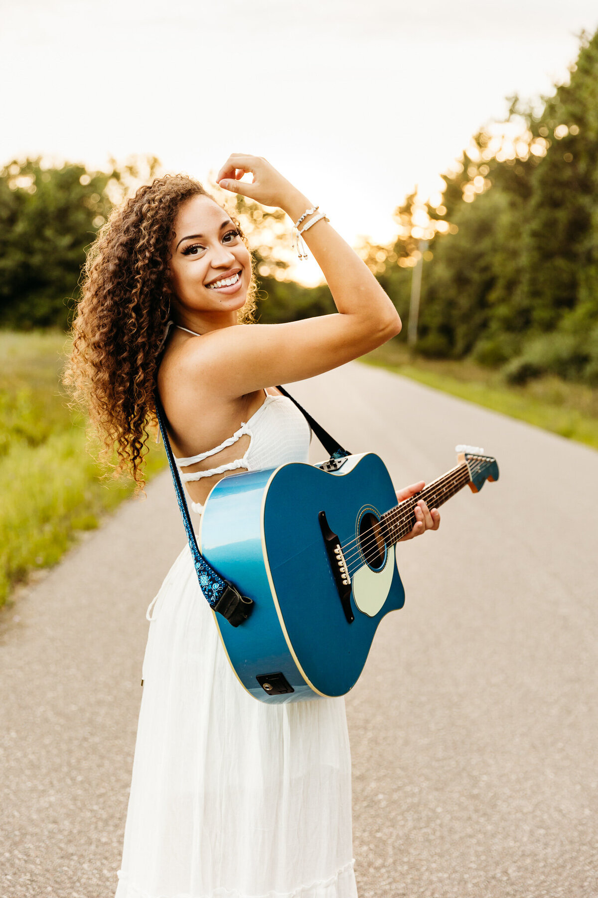 high school senior with curly hair and a white dress smiling while holding a guitar in a road