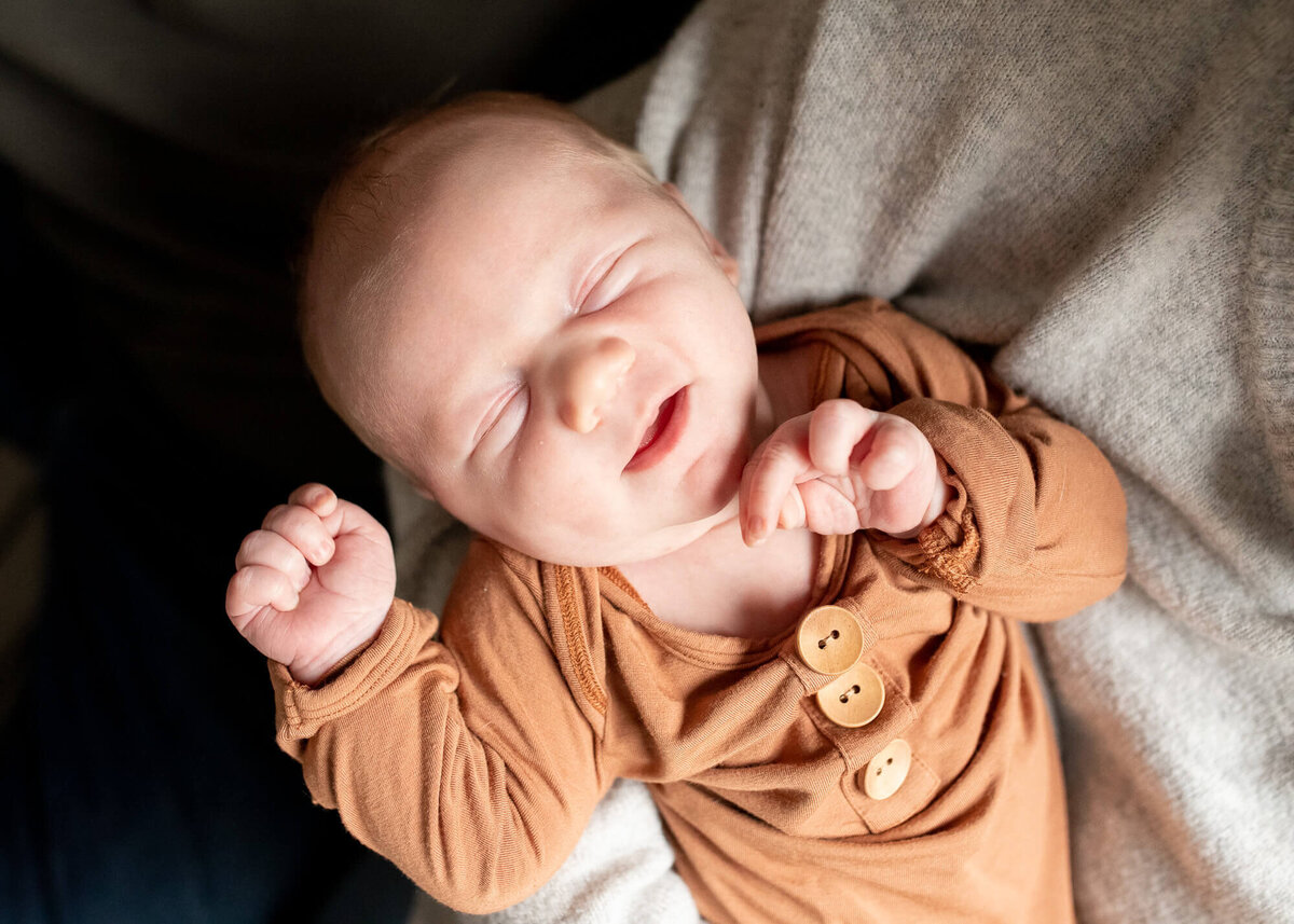 A newborn baby smiling in mothers arms in light brown onesie by Portland newborn photographer Jaime Bugbee.