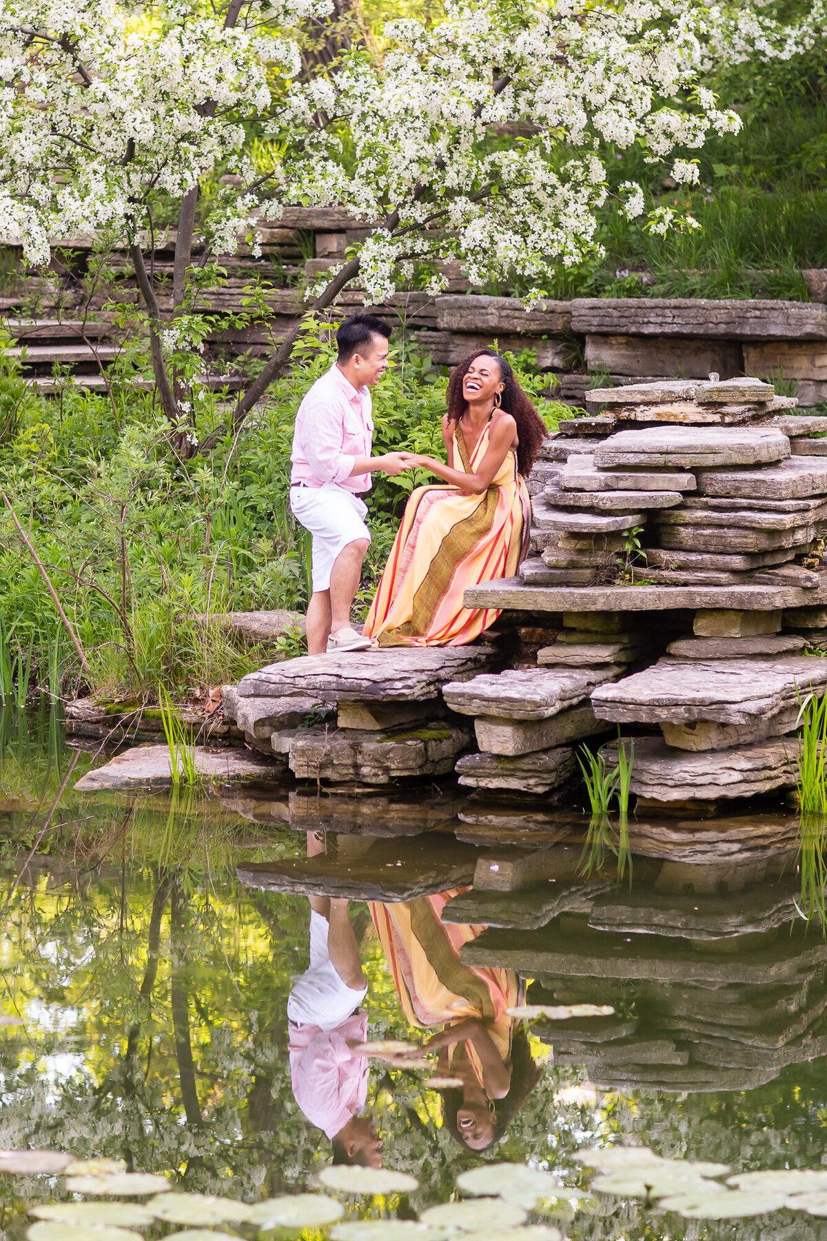 Candid engagement photo of a couple sitting on a rock under a tree in blossom at the Alfred Caldwell Lily Pool in Lincoln Park, Chicago