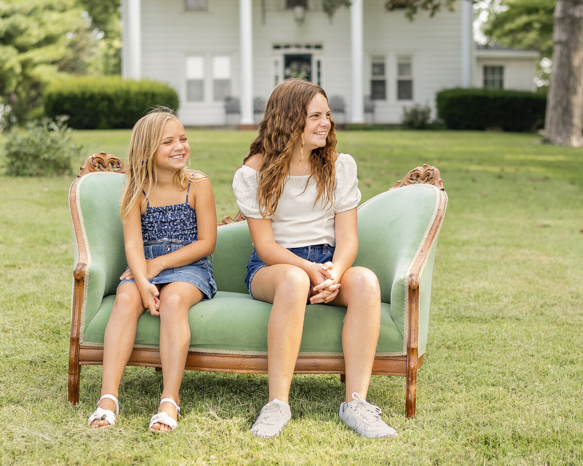 Sisters are sitting on a vintage green couch in the grass with their family two story home in the background, looking away from the camera smiling. Older daughter is wearing blue jean shorts, a white short sleeve shirt, and has long brown hair.  Youngest daughter has a dark blue and white tank top style shirt with a blue jean skirt, and has medium length blonde hair.