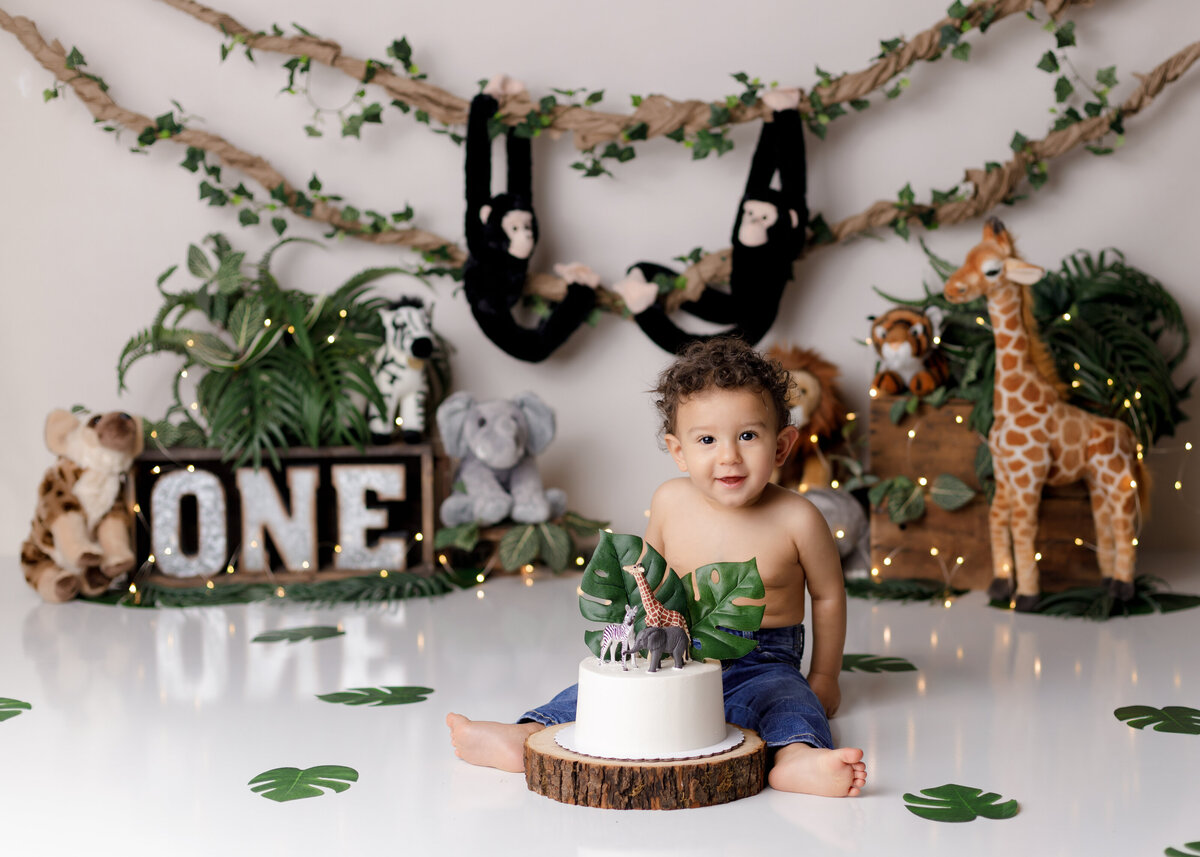 Safari jungle themed cake sash in West Palm Beach and Boca Raton, FL newborn and cake smash photographer studio. Baby boy is wearing overalls and canvas safari hat with cake on his overalls. He is looking at the camera. In the background, there is a jungle themed backdrop, leaves, and stuffed jungle animals.
