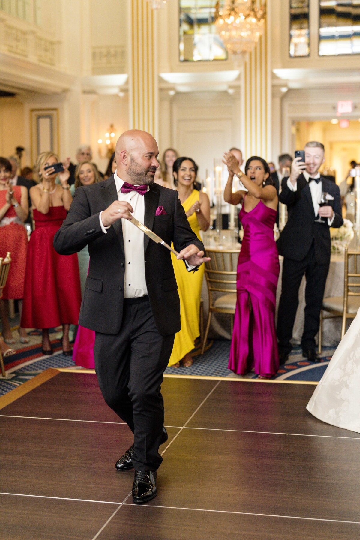 A bald man in a tuxedo is dancing and ribbon twirling at a formal event. Around him, elegant guests in colorful gowns and suits are clapping and cheering, some capturing the moment with their phones. The setting is lavish with chandeliers and gold accents.