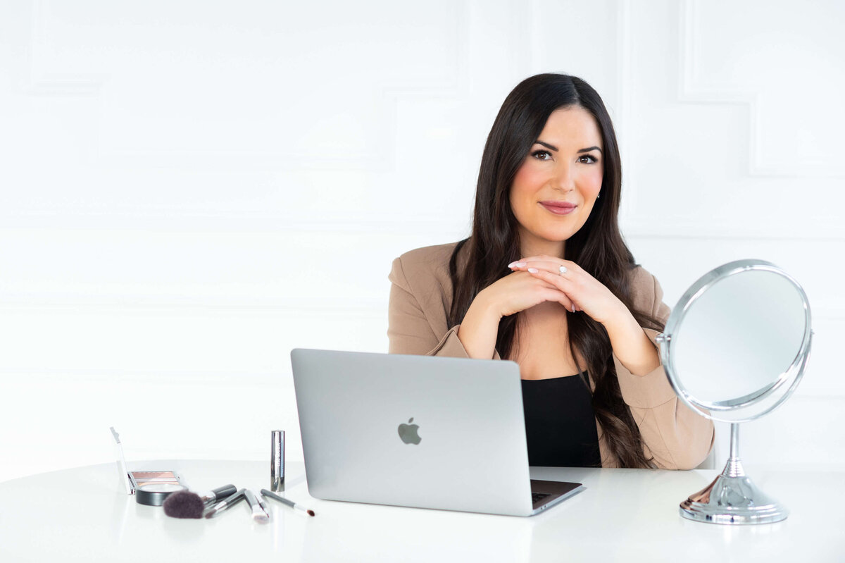 a profile photo a makeup artist sitting at her desk with her laptop, makeup and mirror ready for an online makeup lesson.  Captured indoors at Skye Studio by Ottawa Branding Photographer JEMMN Photography COMMERCIAL