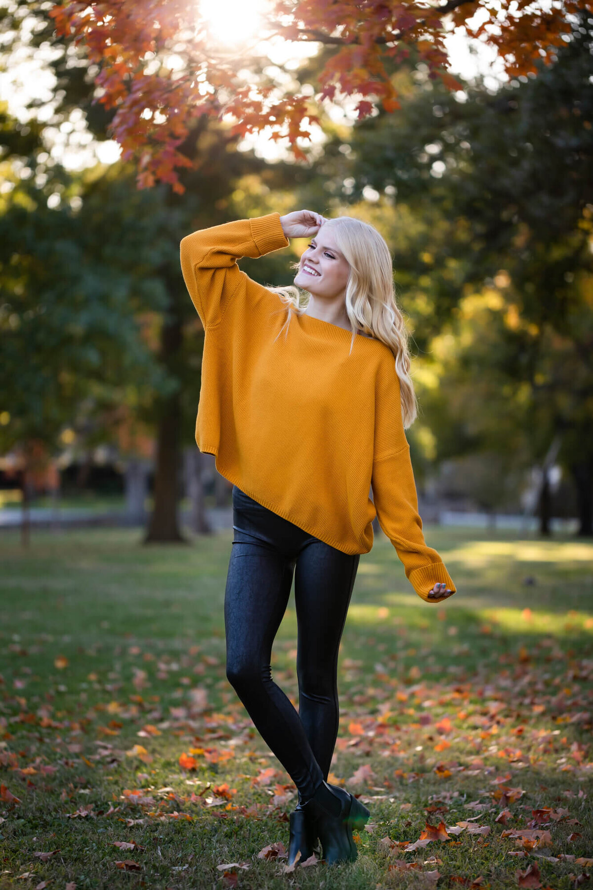 A beautiful blonde teen girl posing in the Fall sun wearing a dark yellow sweater and black leather leggings. Captured by Springfield, MO senior photographer Dynae Levingston.