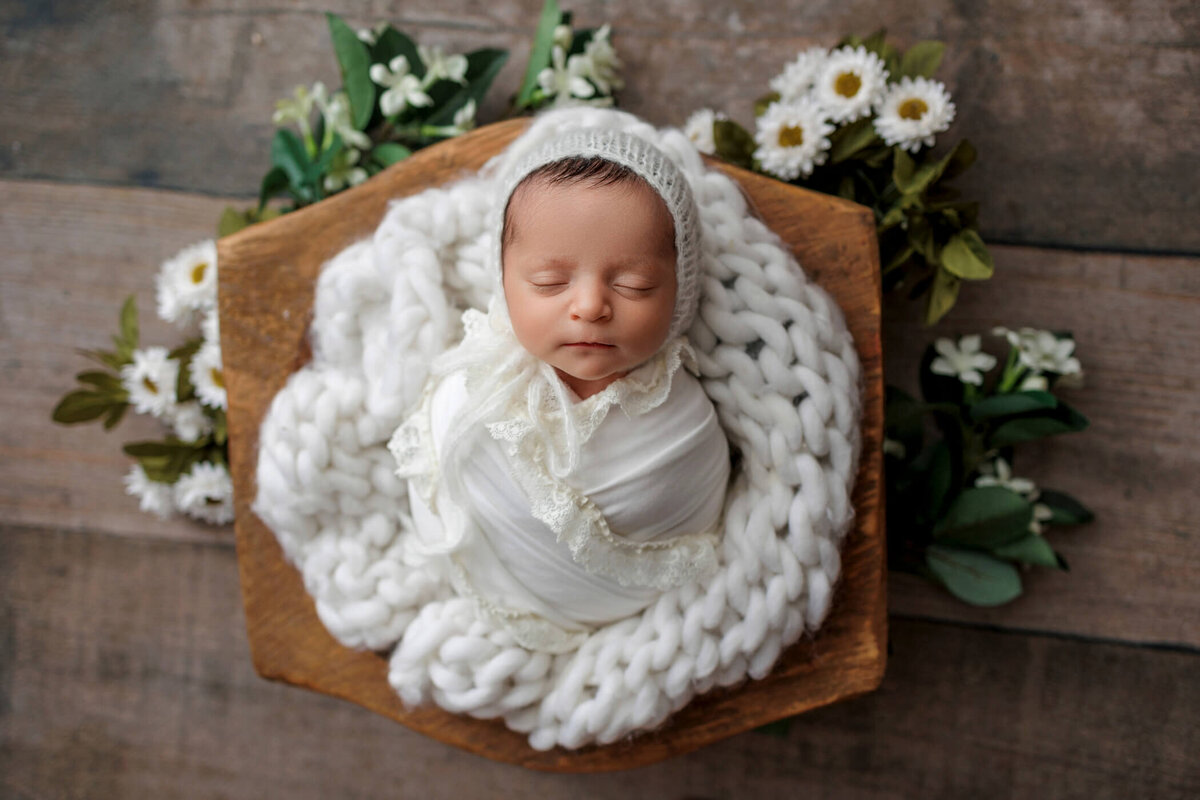 Newborn baby wrapped in a white swaddle, wearing a white knit bonnet, sleeping on a chunky white knit blanket surrounded by white flowers, set against a rustic wooden background.