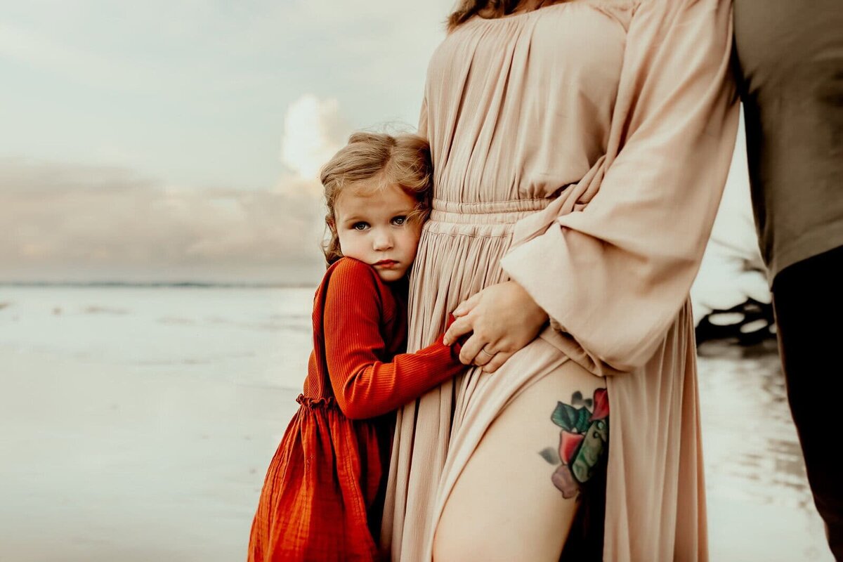 Child in red dress hugs adult's leg near the beach. Adult wears a beige dress with a leg tattoo. Overcast sky and water in the background.