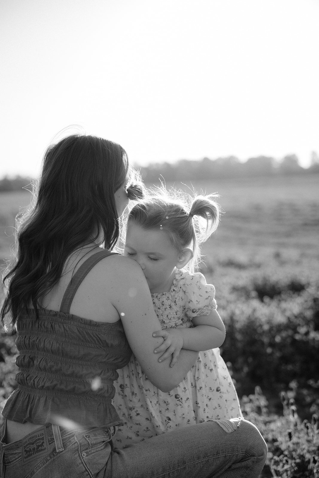 Daughter kisses her mothers shoulder as mother hugs her daughter at sunset.