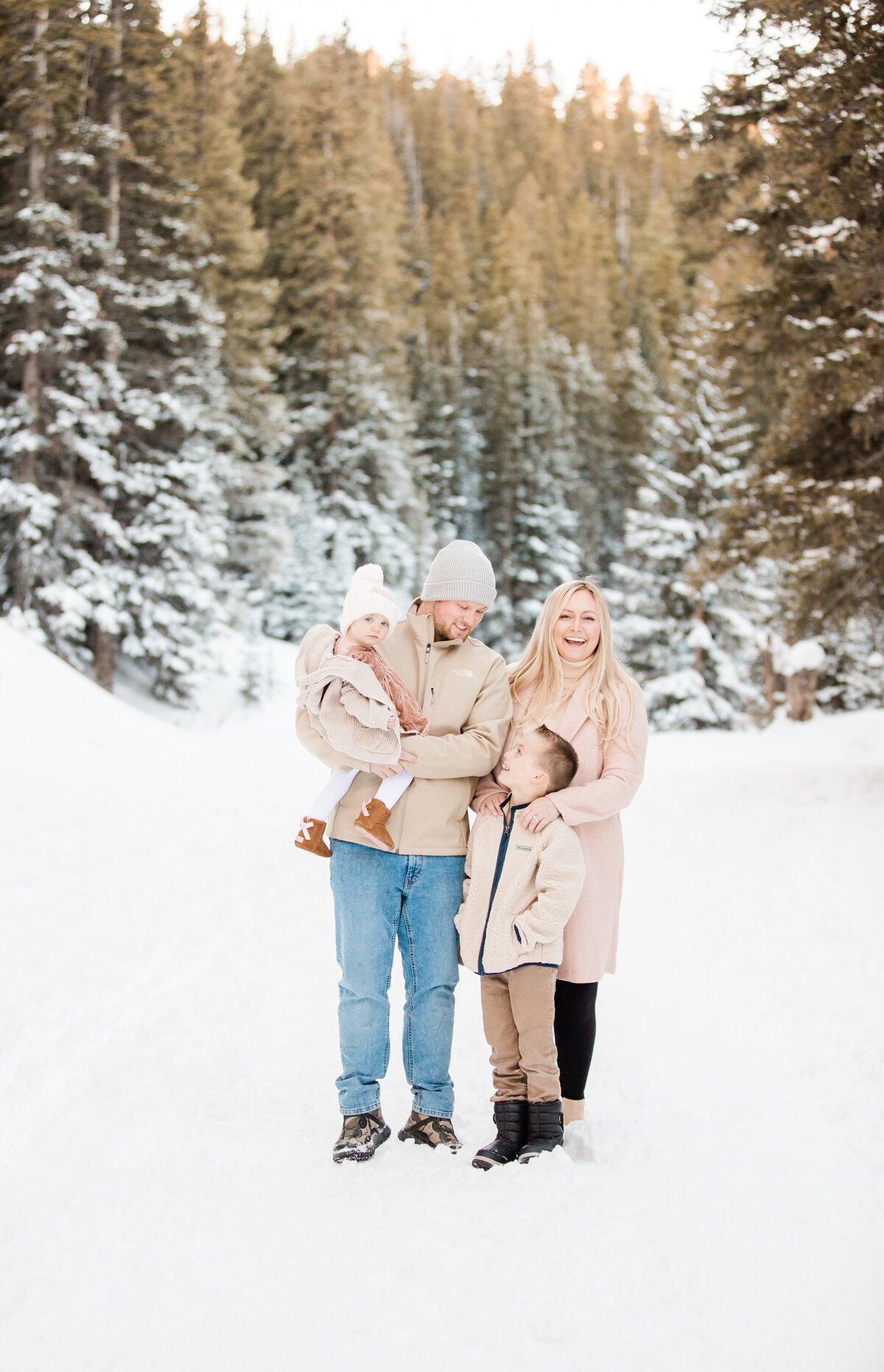 A family of four stands close together for a photo. A winter scene of snow covered pine trees are behind them