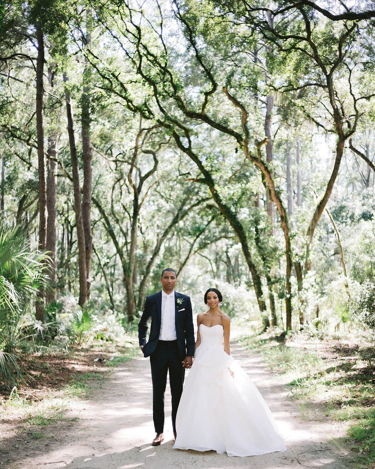 The bride and groom are standing holding hands amongst the trees in Montage at Palmetto Bluff. Destination wedding image by Jenny Fu Studio