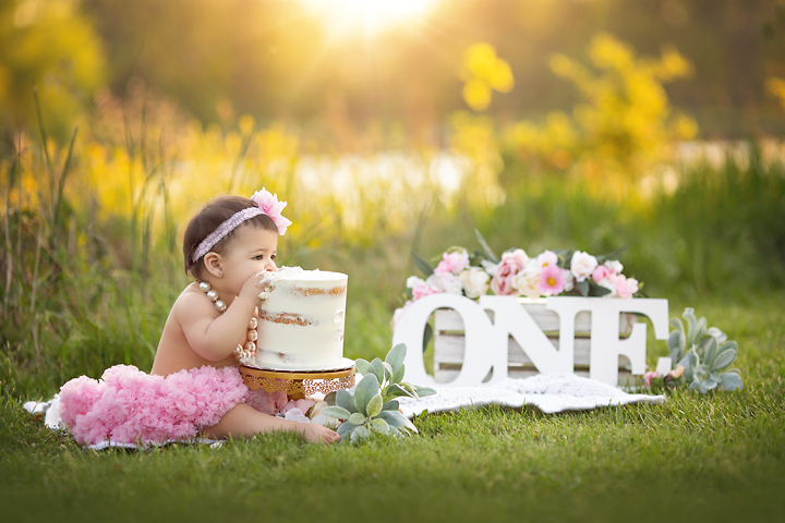 A young toddler girl takes a bite out of a cake while sitting in a grassy lawn on a pond for her first birthday