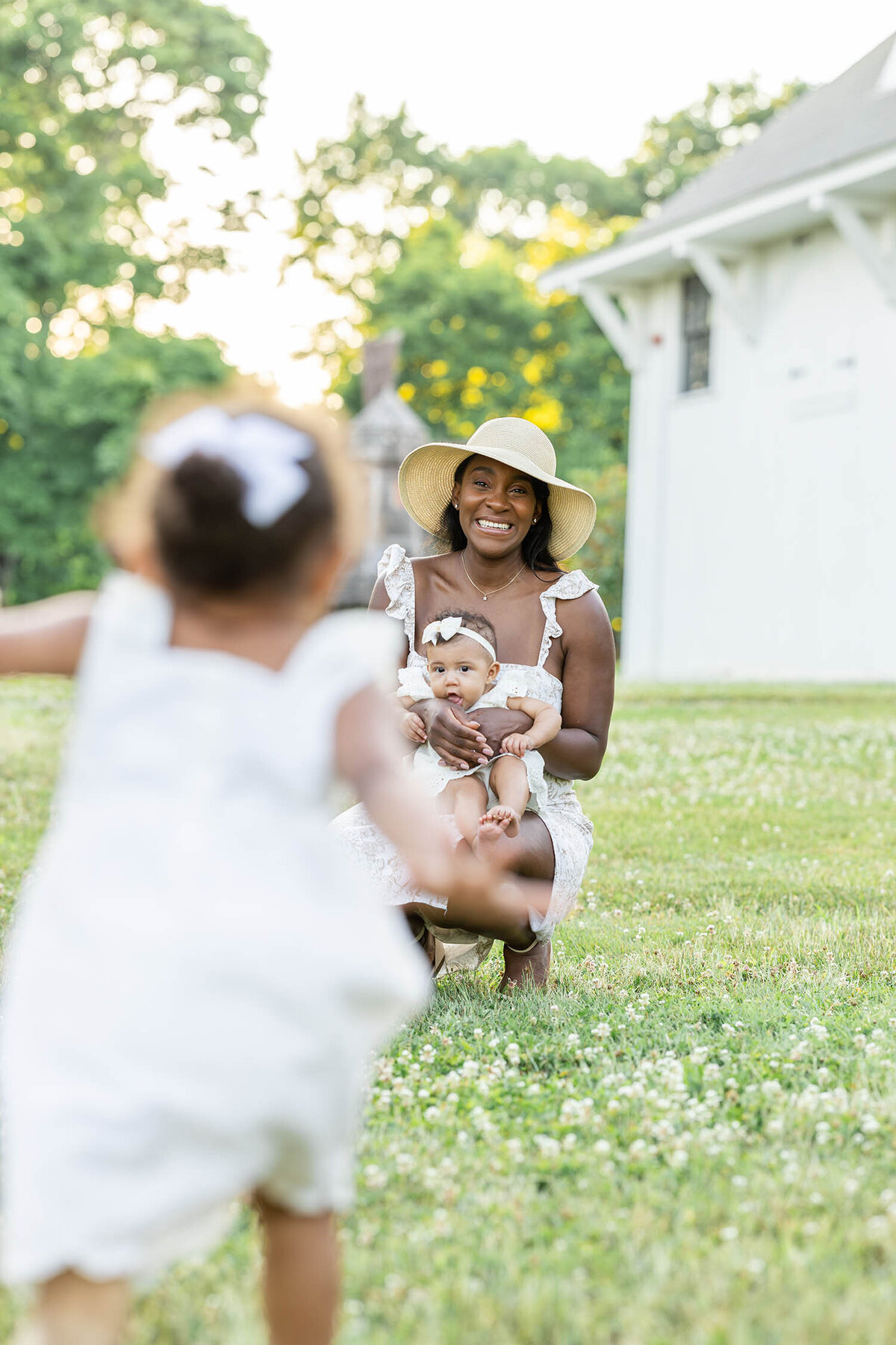 Toddler girl is running towards woman who is kneeling down and holding baby on her lap in a clover field of grass 