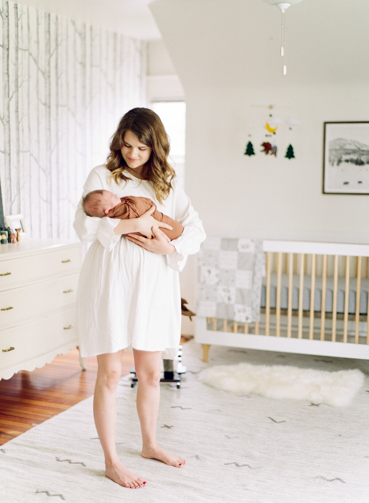 Mom stands while rocking her newborn son in his white nursery in Raleigh NC newborn photo session. Photographed by Newborn Photographers Raleigh A.J. Dunlap Photography.